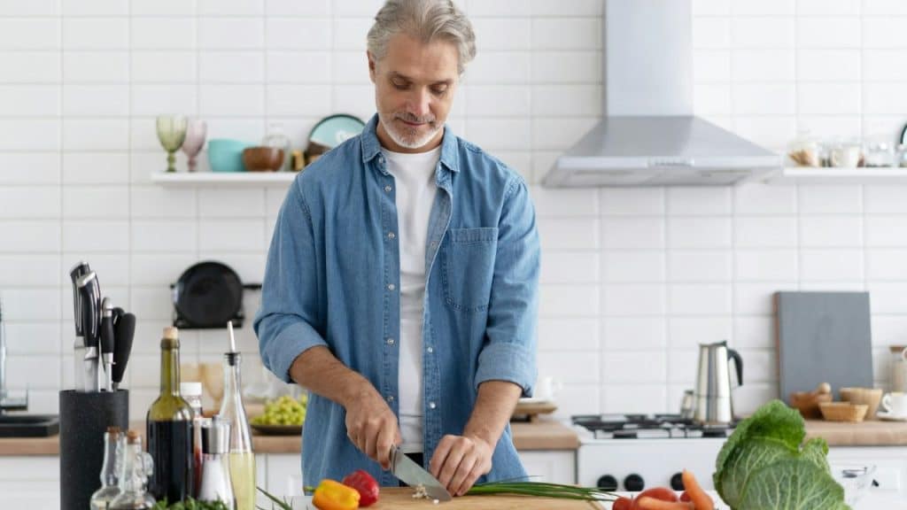 A gray-haired man in a denim shirt chops vegetables on a cutting board in a kitchen.