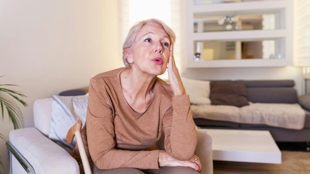 An older woman sitting on a couch with her hand on her face, looking worried and tired.