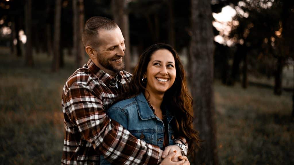 A smiling man embraces a woman from behind in a forest setting.