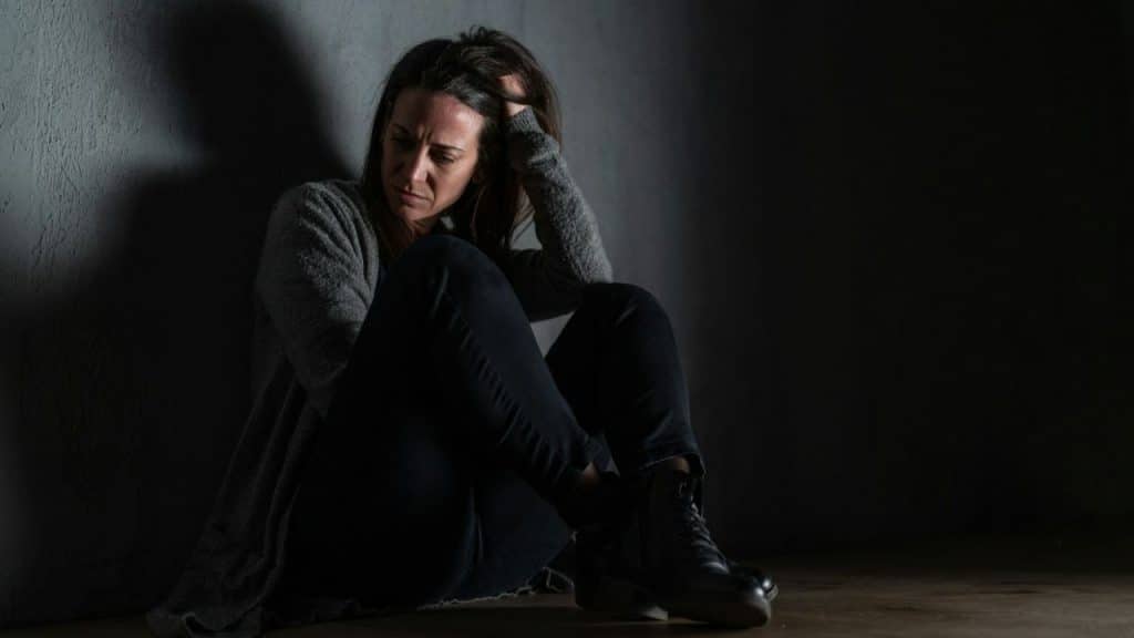 A distraught woman sits alone in the dark, hugging her knees with her head down.