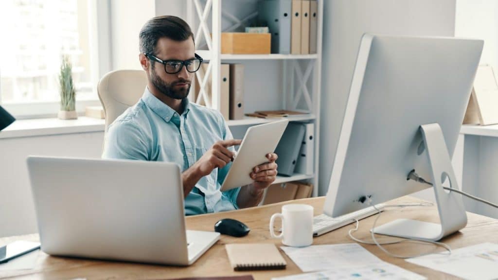 A man in a light blue shirt and glasses working on a tablet at a desk.