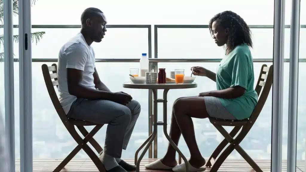 A man and woman sit opposite each other on a balcony, having a silent meal.