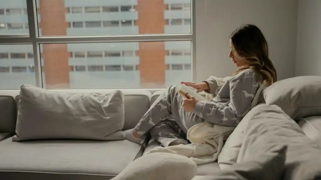 Woman in pajamas reading a book while relaxing on a couch by a large window.