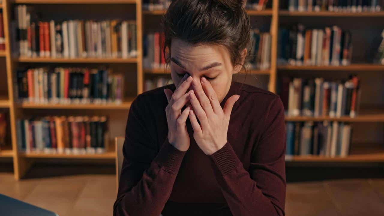 A person sitting in a library rubbing their eyes in frustration or fatigue.