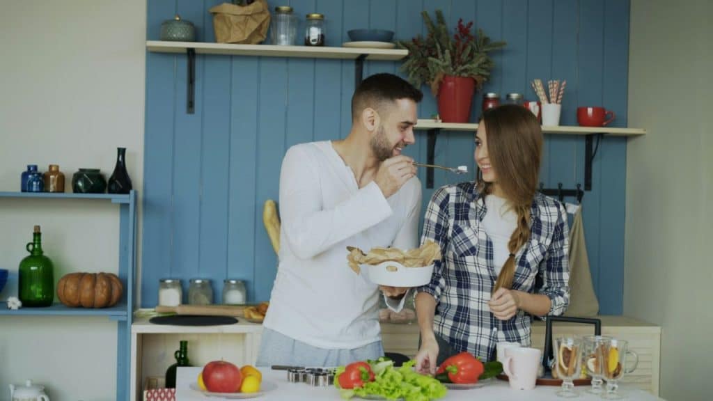 Smiling man feeding a woman with a chopstick in a bright kitchen with blue walls.