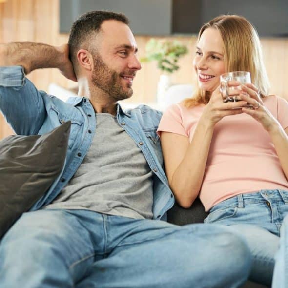 A couple smiling and relaxing on a sofa; the woman holds a glass of water.