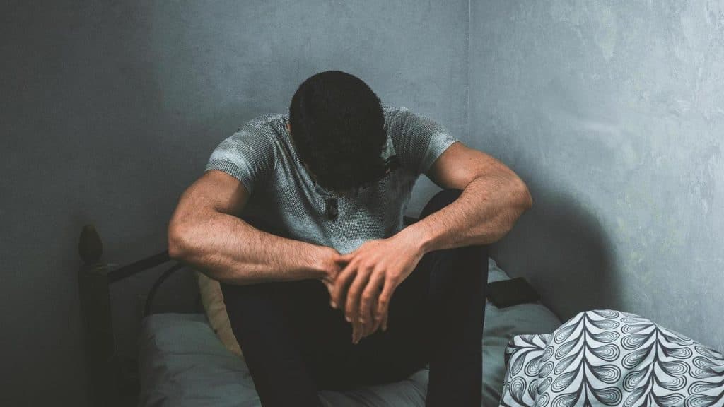 A man sitting on a bed with his head bowed and arms resting on his knees.