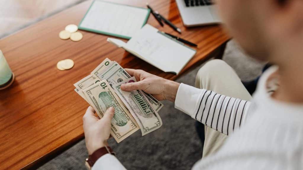 A person counting US dollar bills at a wooden table with notebooks and coins.