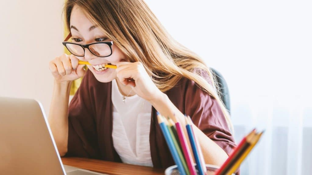 A woman biting a pencil while sitting on a chair in front of a computer during the daytime.