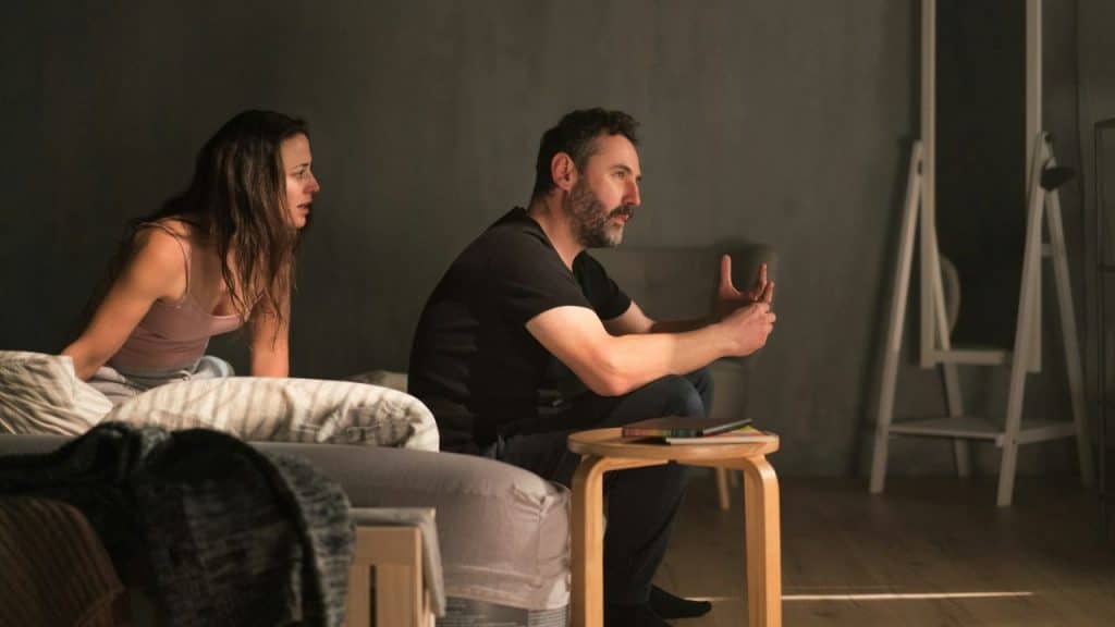 Man and woman sitting indoors in a dim room with the man gesturing while talking.