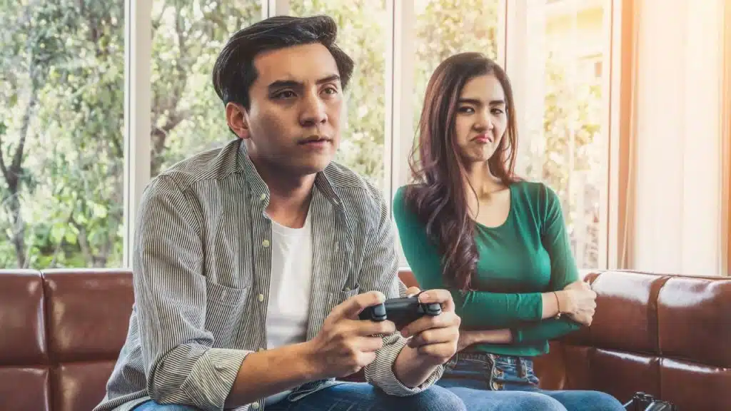 A man intently plays a video game with a controller while a woman next to him looks disgruntled.