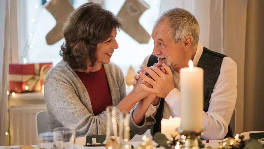 Elderly man and woman smiling, holding hands across a table with candles.