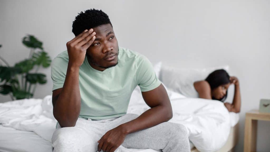 A man looking stressed while sitting on a bed with a woman lying down, turned away in the background.