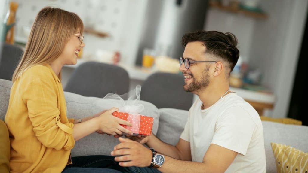 Smiling couple on a couch; woman gives a man a small red gift box.