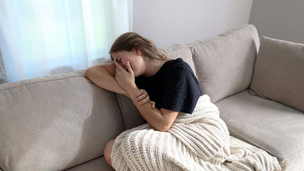 A tearful woman wrapped in a blanket on a couch, covering her face with her hand.
