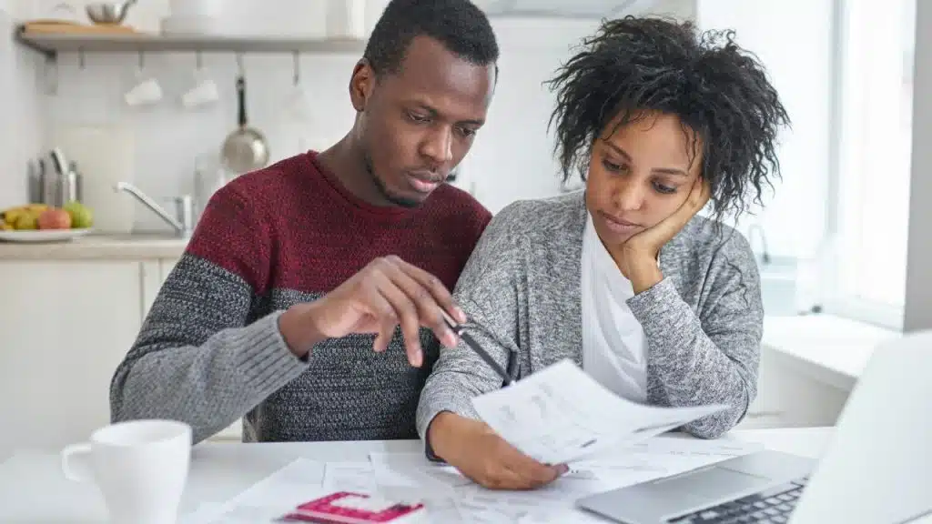 A man and a woman sit at a table reviewing papers in a brightly lit kitchen.