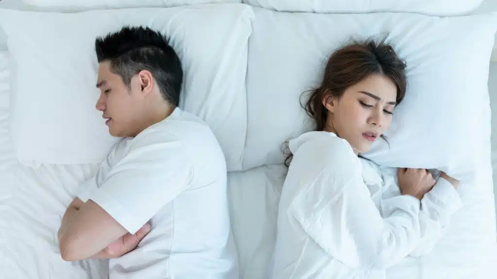 An overhead view of an Asian couple lying in bed, turned away from each other in disagreement, with the man's arms crossed.