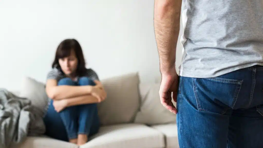 A blurry woman sits sadly on a couch as a man's back, wearing a gray shirt, faces away.
