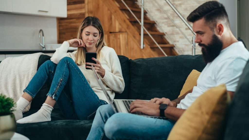 Woman sitting on a couch, looking at her phone next to a man with a laptop.