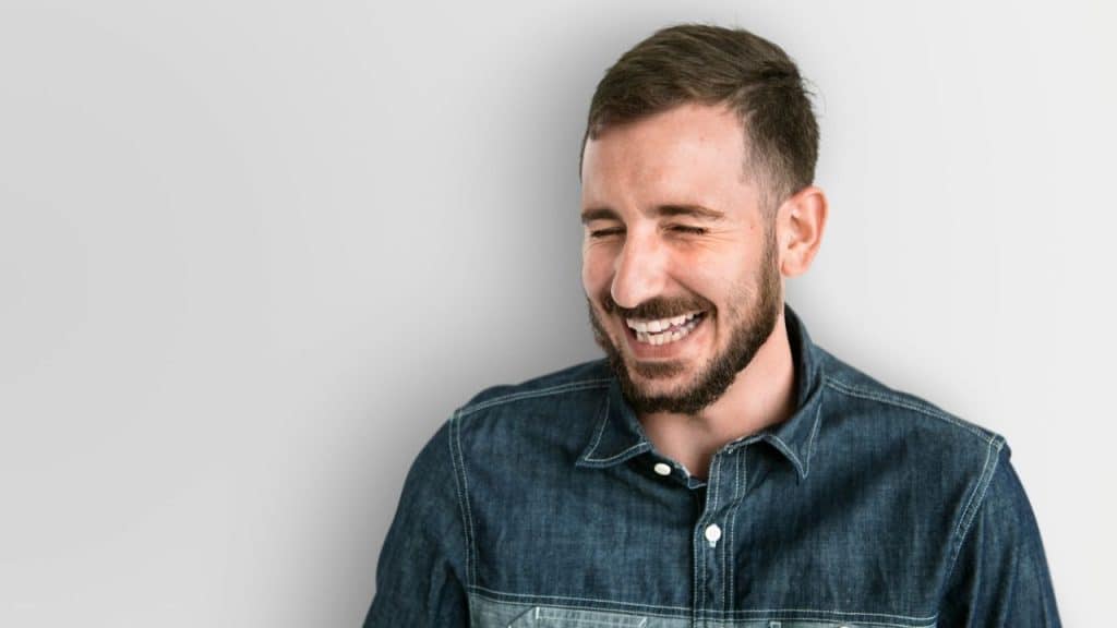Smiling man with beard and denim shirt laughing with eyes closed against a light wall.