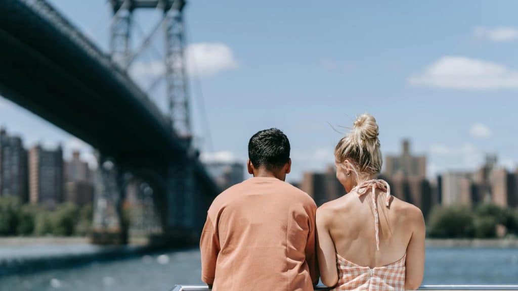 A couple sitting by the water with a bridge in the background.