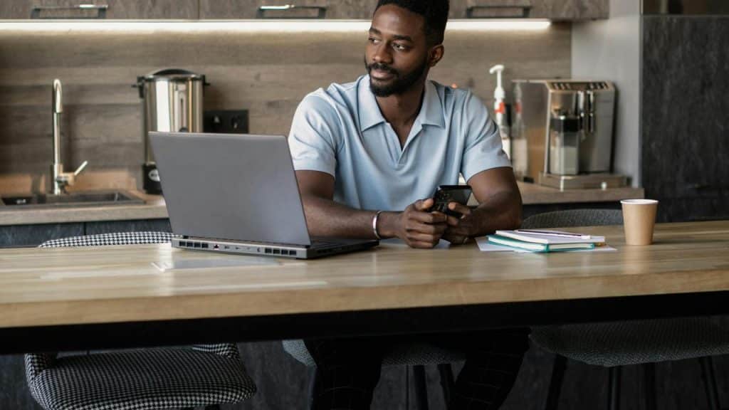 A man sitting at a kitchen table using a laptop and holding a phone.