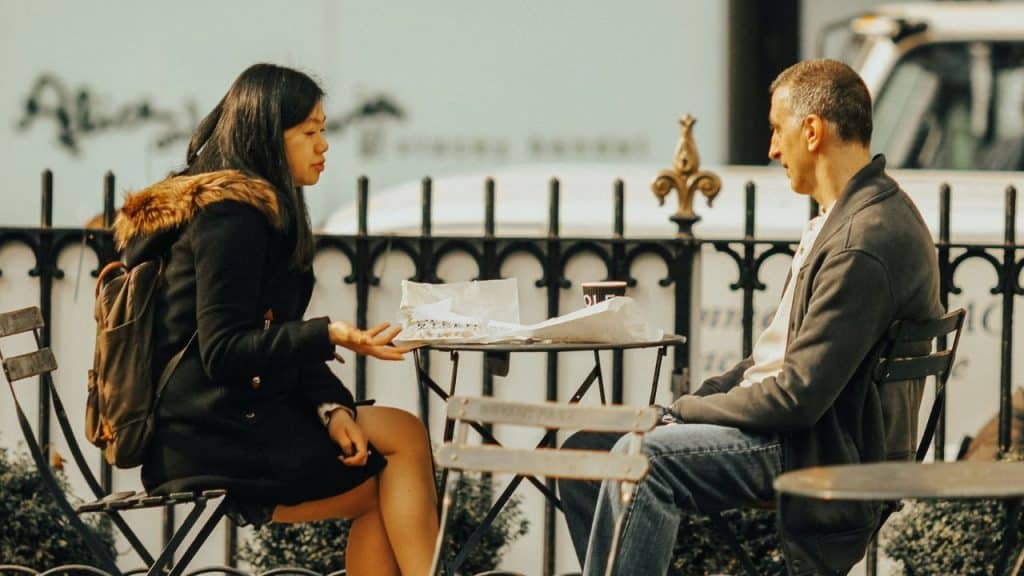 A man and a woman sit at an outdoor table with food, engaged in conversation.