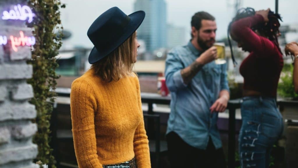 A woman in a yellow sweater and blue hat looks toward a man holding a drink.