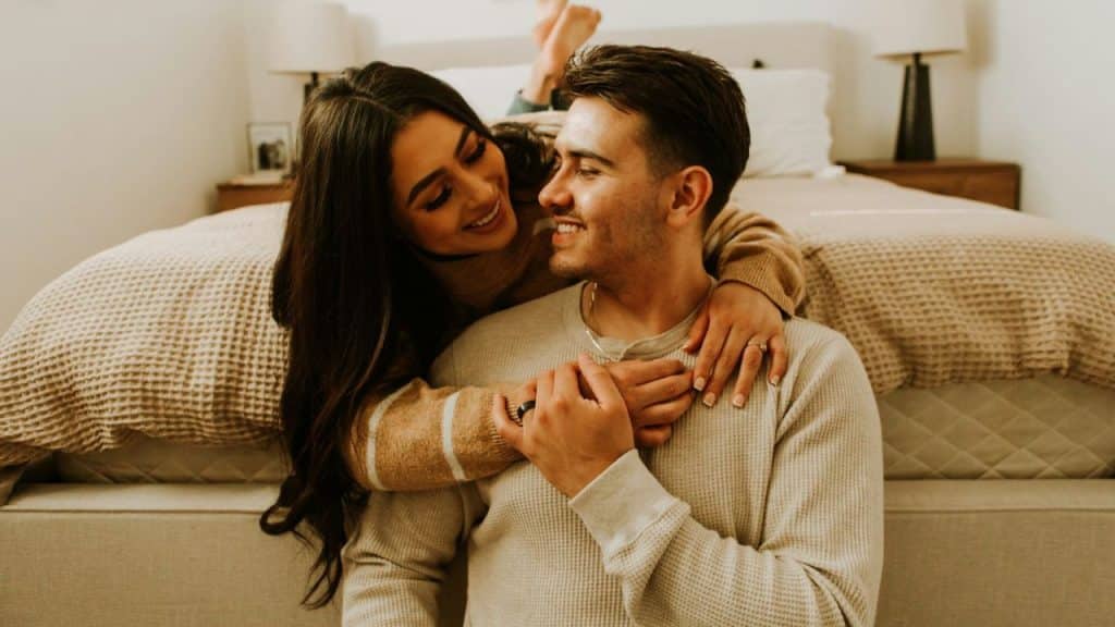 A woman leans over a man’s shoulders while they look at each other and smile.
