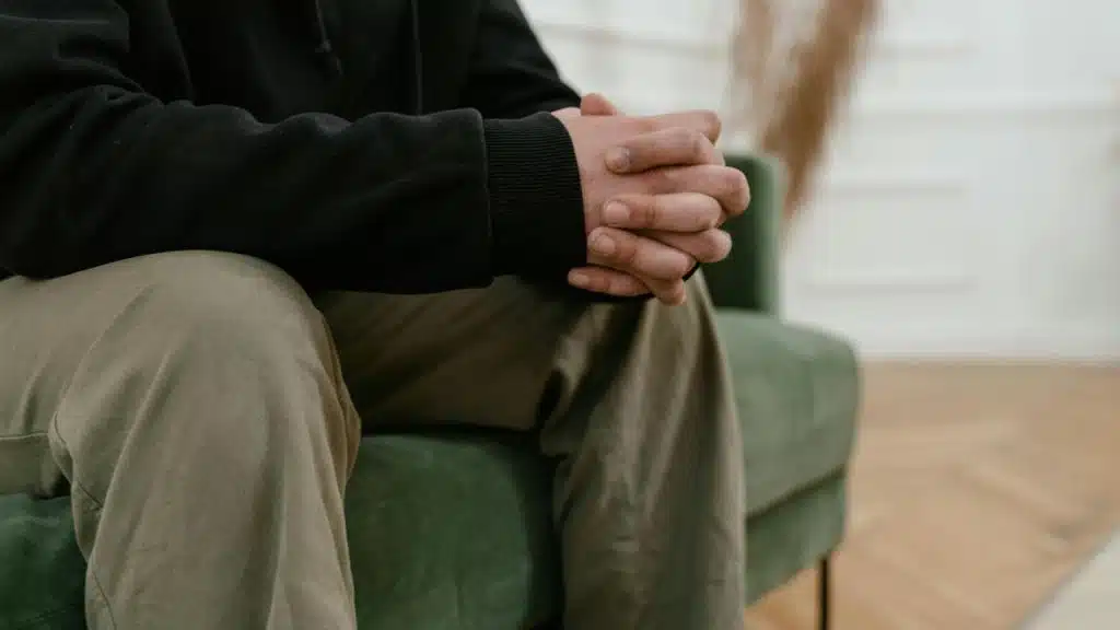 A person sitting on a couch with hands clasped, appearing thoughtful.