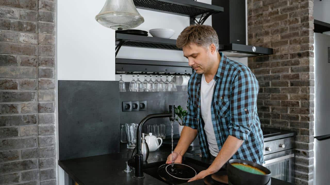 A man in a checkered shirt washes dishes at a kitchen sink.