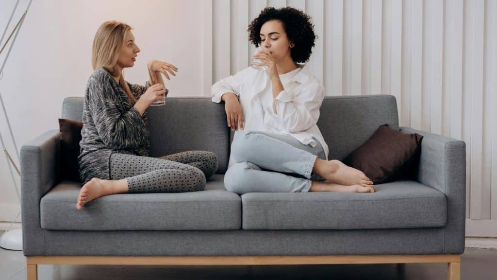 A pair of women sitting on a sofa, talking and drinking water.