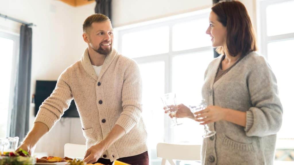 A man and woman in cozy sweaters talk while setting a table.