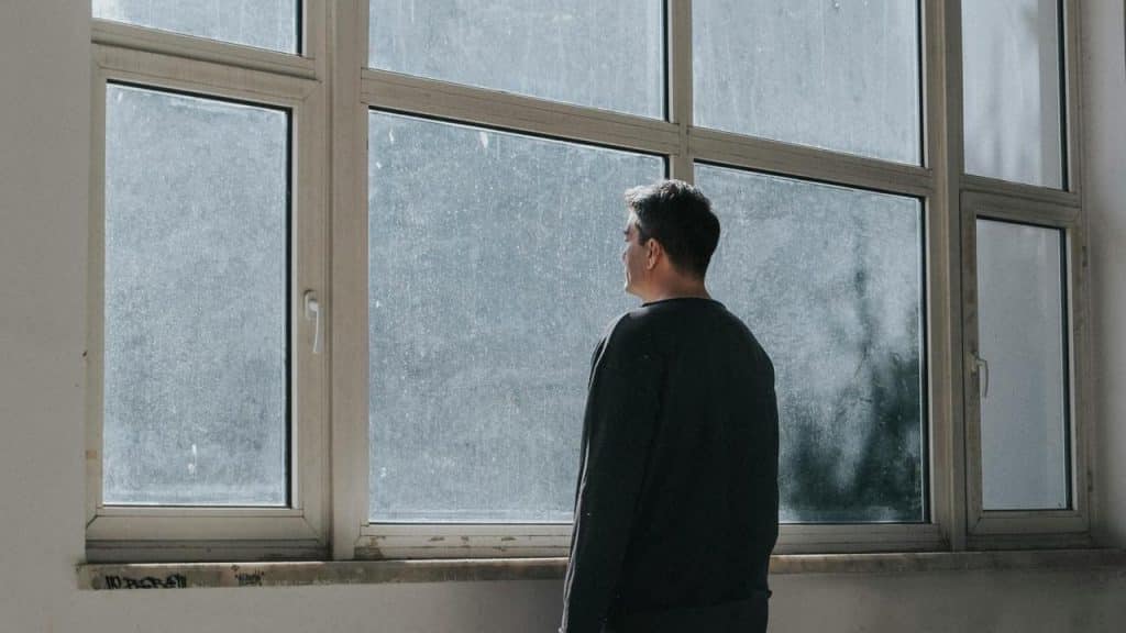 A man standing indoors facing a large window, looking outside in soft daylight.
