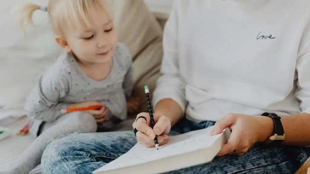 A toddler watching an adult draw in a sketchbook while sitting together on a couch.