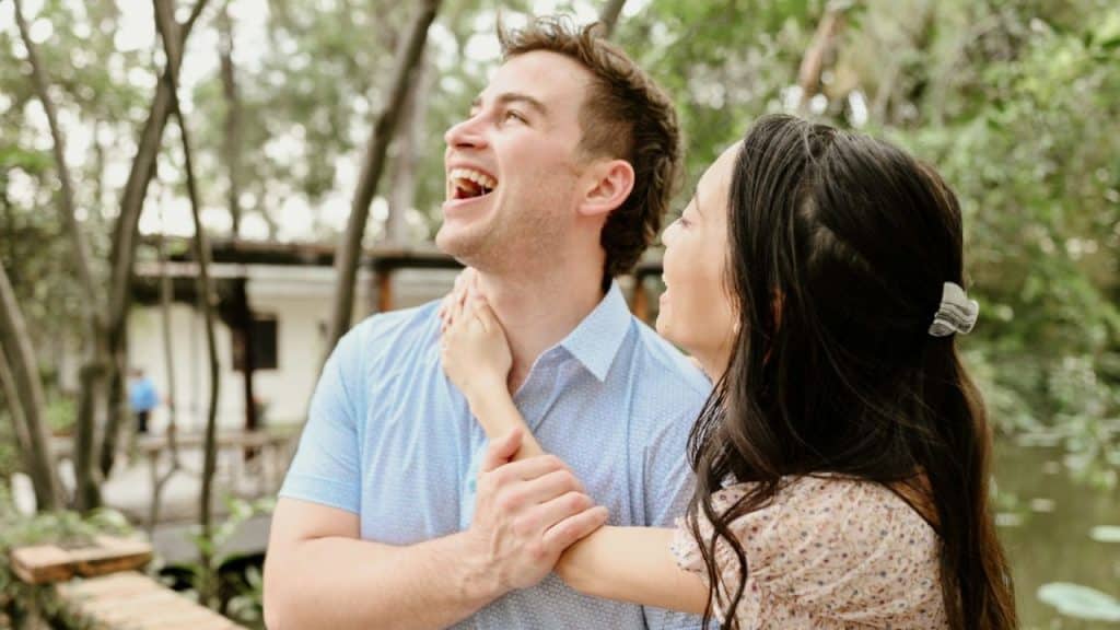 A man and woman laugh together while standing in a lush, green outdoor setting.