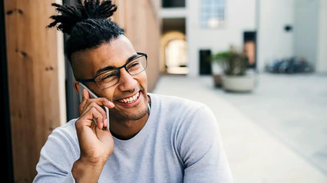 A man wearing glasses smiling while talking on his phone outdoors.