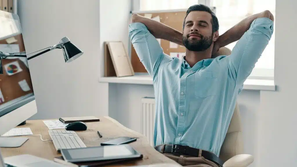 A bearded man leans back in an office chair with his hands behind his head.