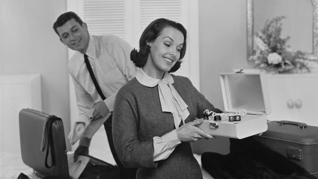 A man packing his suitcase while his wife is holding a small box.