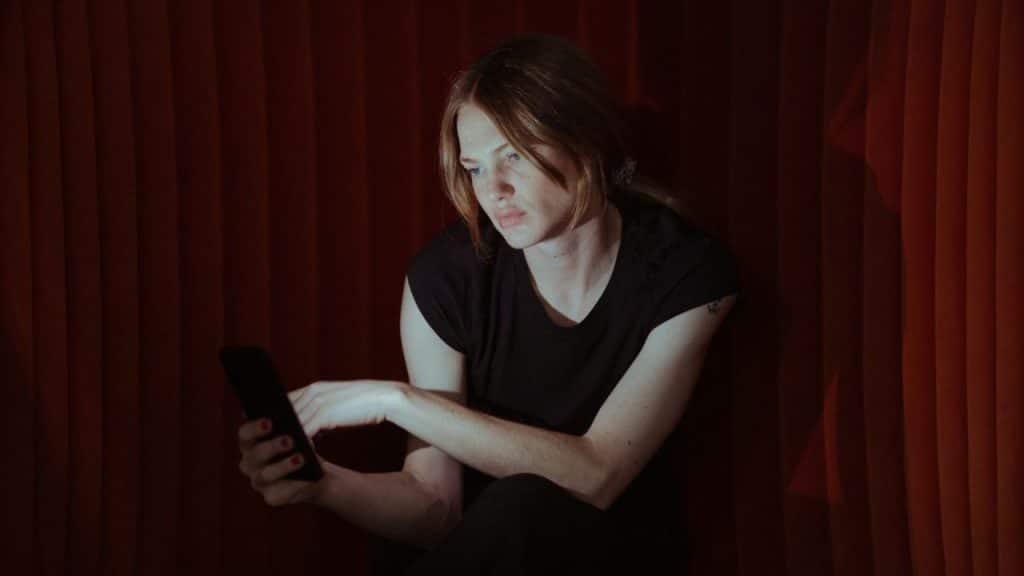 Woman in a black shirt looking at her phone against a dimly lit red curtain background.