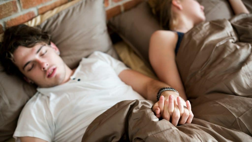 A couple sleeping in bed, holding hands under the covers.