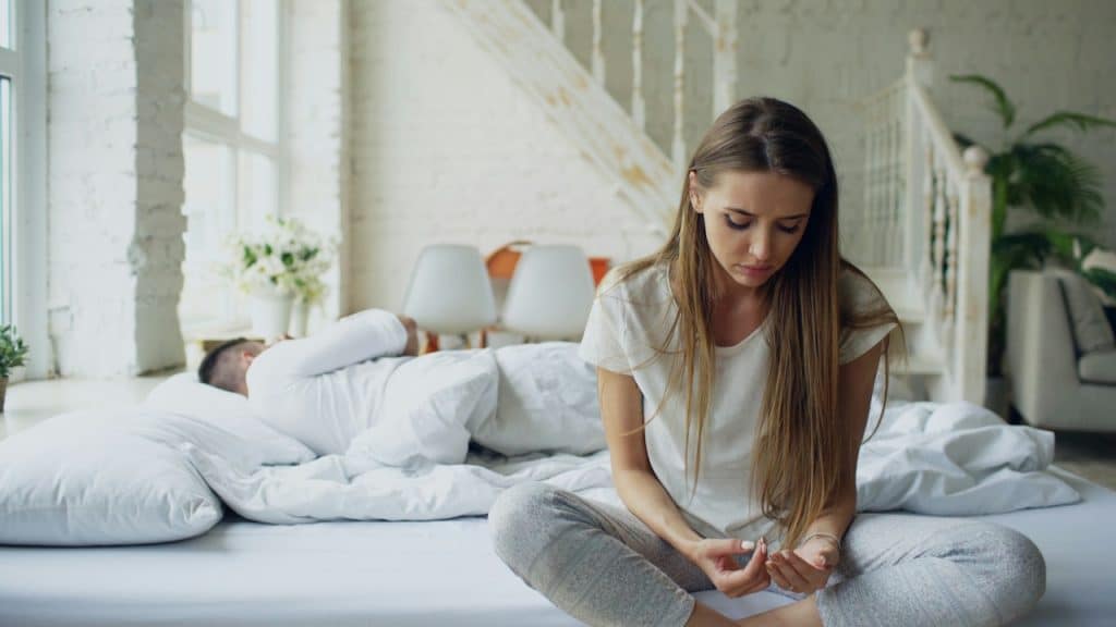 A woman is sitting cross-legged on a large white bed, looking down with a somber expression, while a man is lying asleep under the covers behind her.