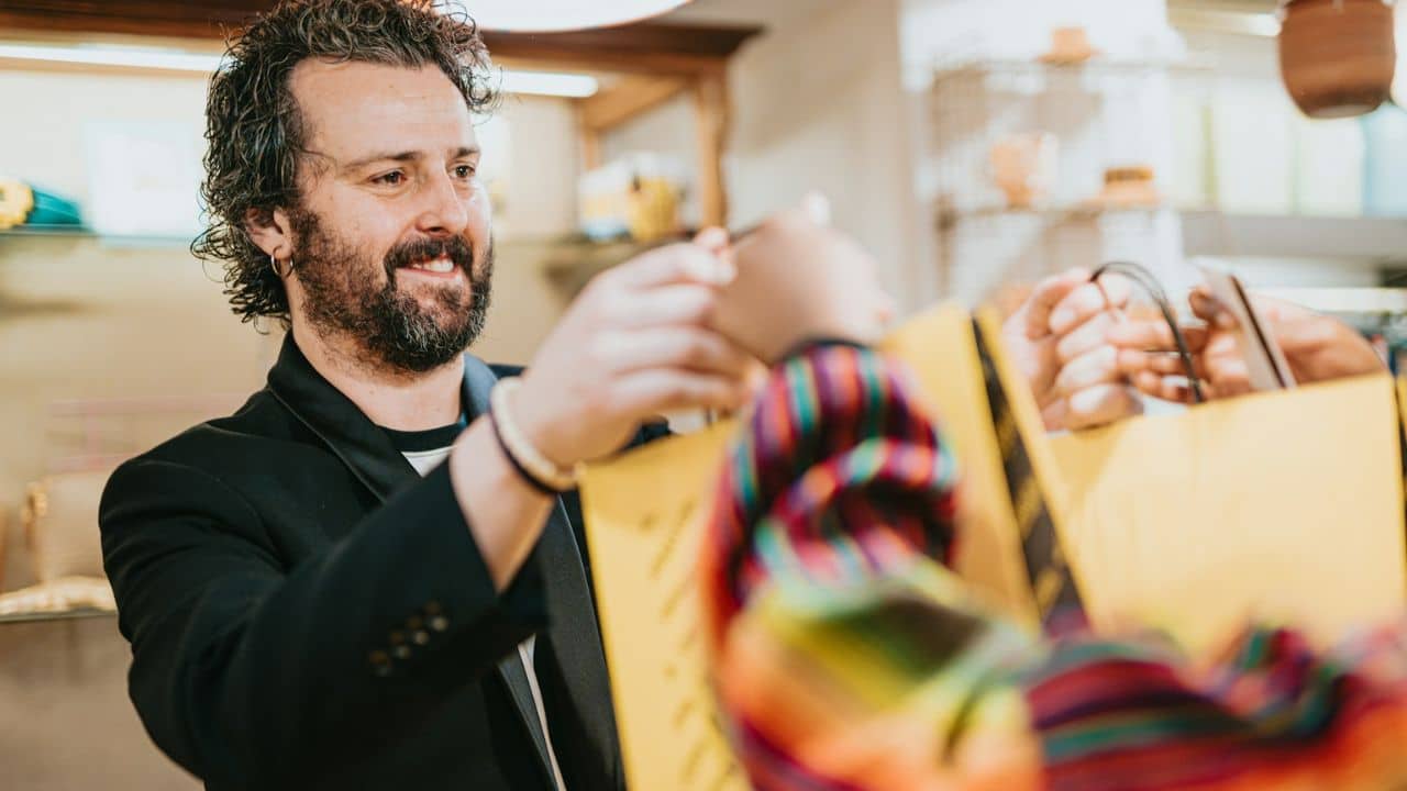 Bearded man smiling while handing yellow shopping bags to an out-of-focus person.