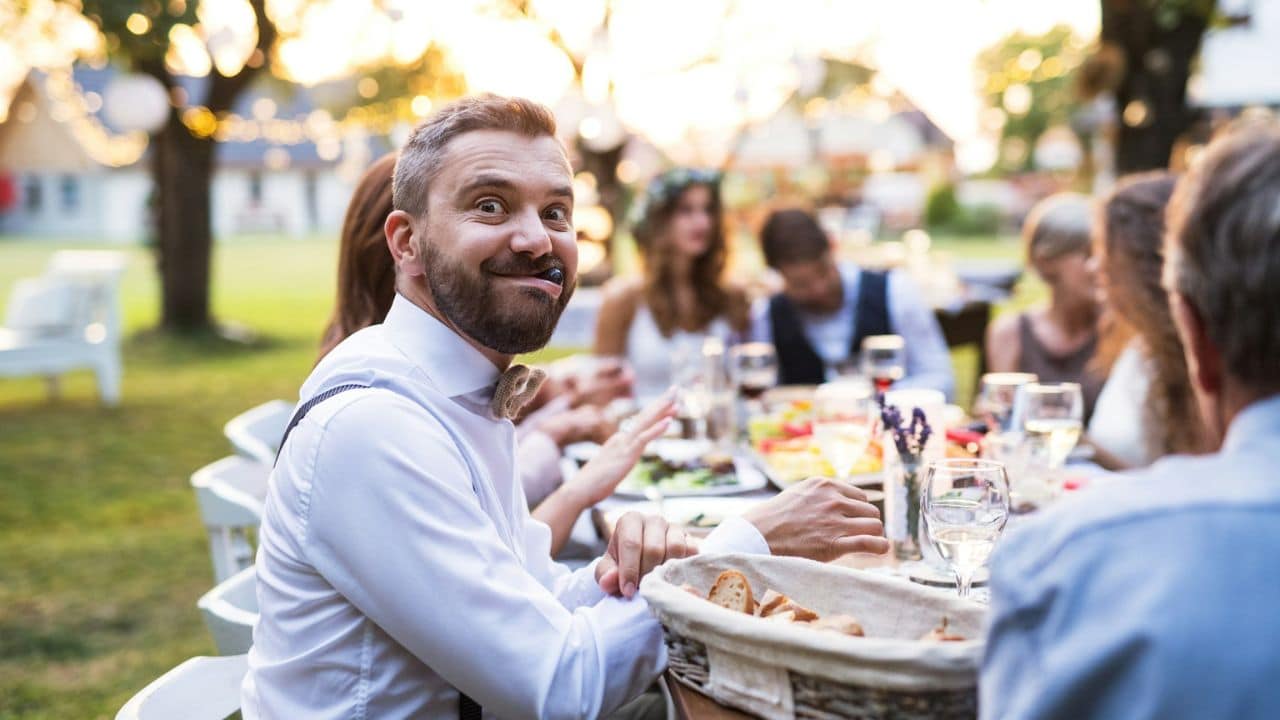 Man with a beard and bowtie making a silly face while sitting at an outdoor dinner table.