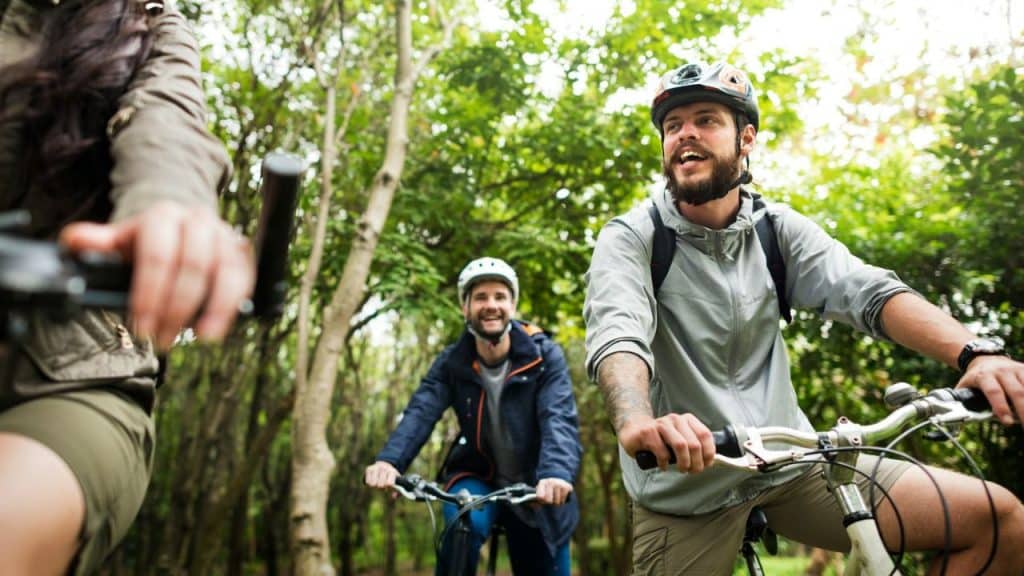 Three people wearing helmets are smiling while riding bicycles on a wooded trail.