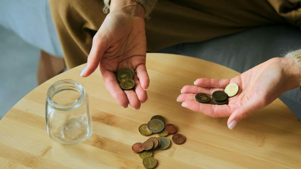 A person's hands holding and counting a small pile of coins next to an empty jar.