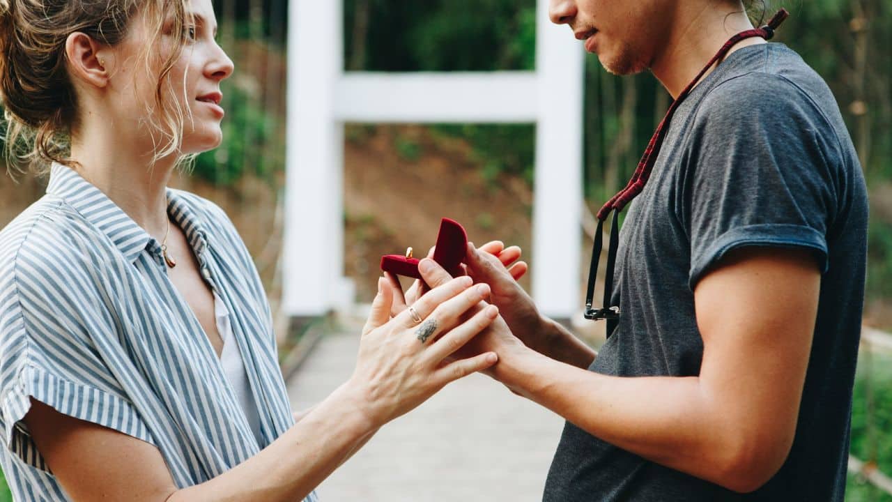 A man is proposing to a woman outdoors, holding out a red ring box.