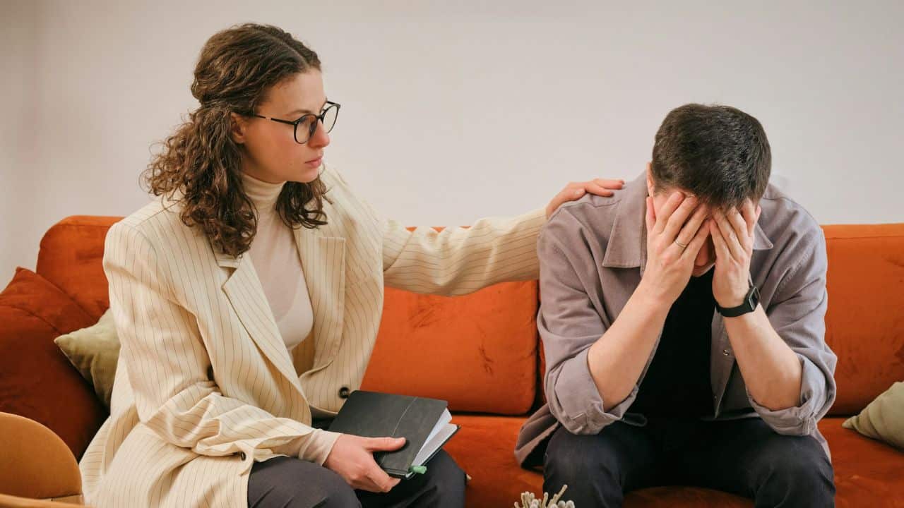 A woman in glasses and a blazer consoles a distressed man holding his head on an orange couch.