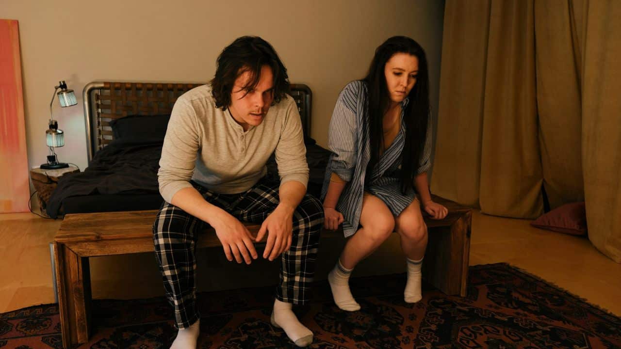 Couple sitting apart on a wooden bench at the foot of a bed, looking downcast.