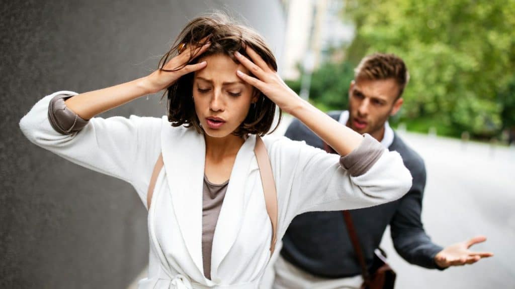 Woman holding her head in distress while a man gestures aggressively behind her outdoors.